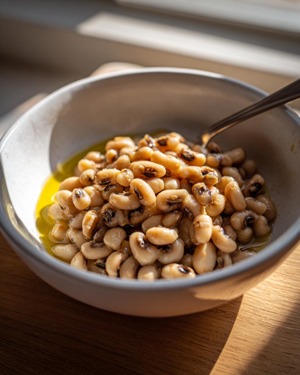 A close-up of a bowl filled with creamy black eyed peas, glistening in a light broth, with a spoon resting inside.