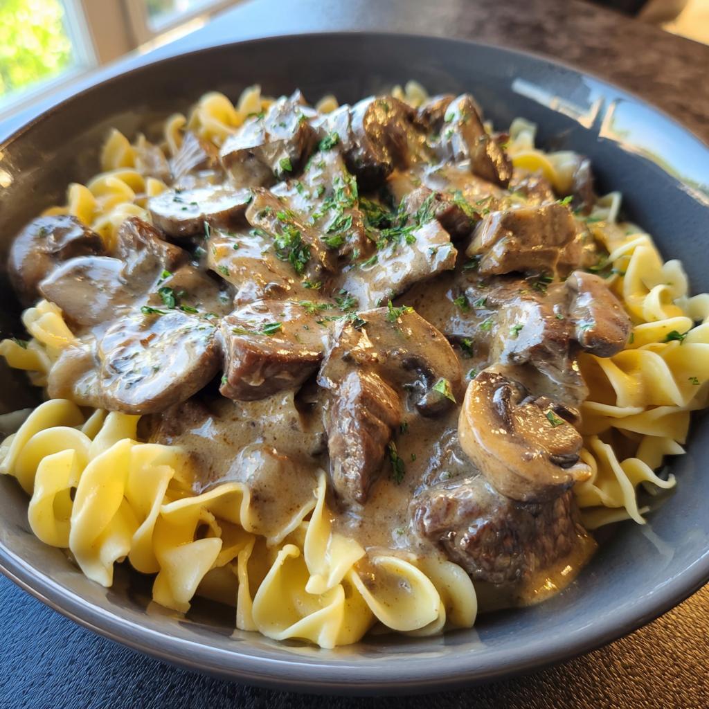 Bowl of creamy beef stroganoff with mushrooms served over egg noodles garnished with herbs.