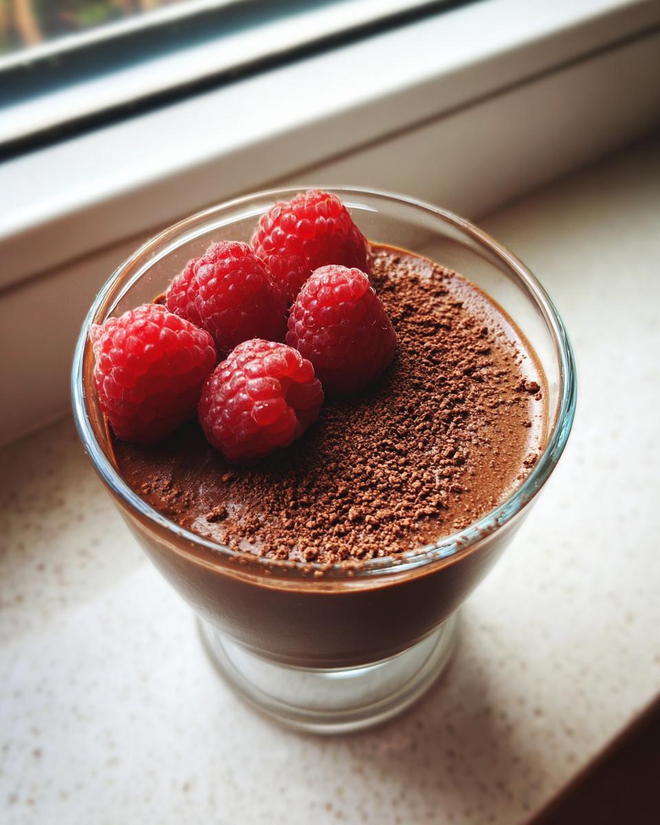 Glass filled with healthy chocolate avocado mousse dessert topped with fresh raspberries and cocoa powder.