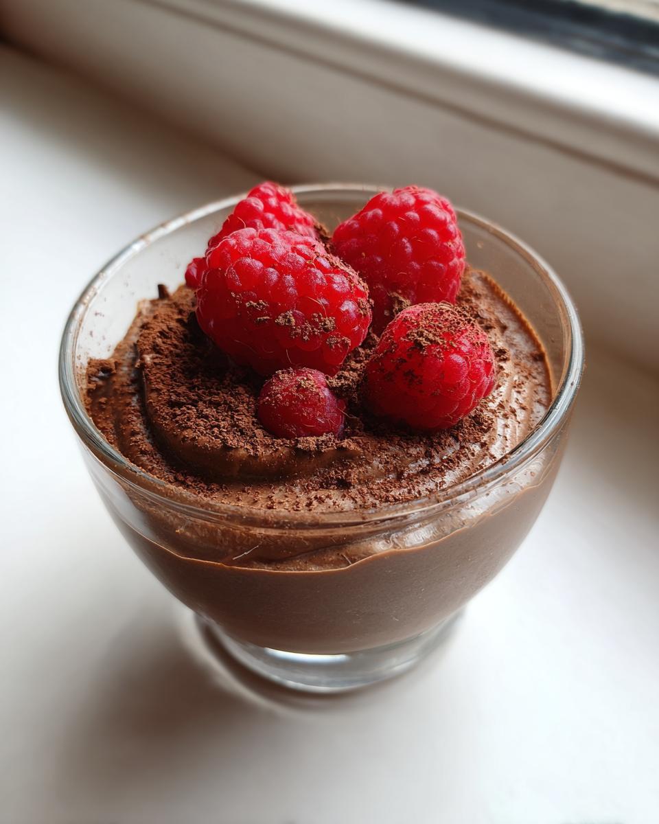 Glass of healthy chocolate avocado mousse dessert topped with fresh raspberries and cocoa powder.