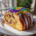 Slice of king cake with white icing and purple, green, and gold beads on top.
