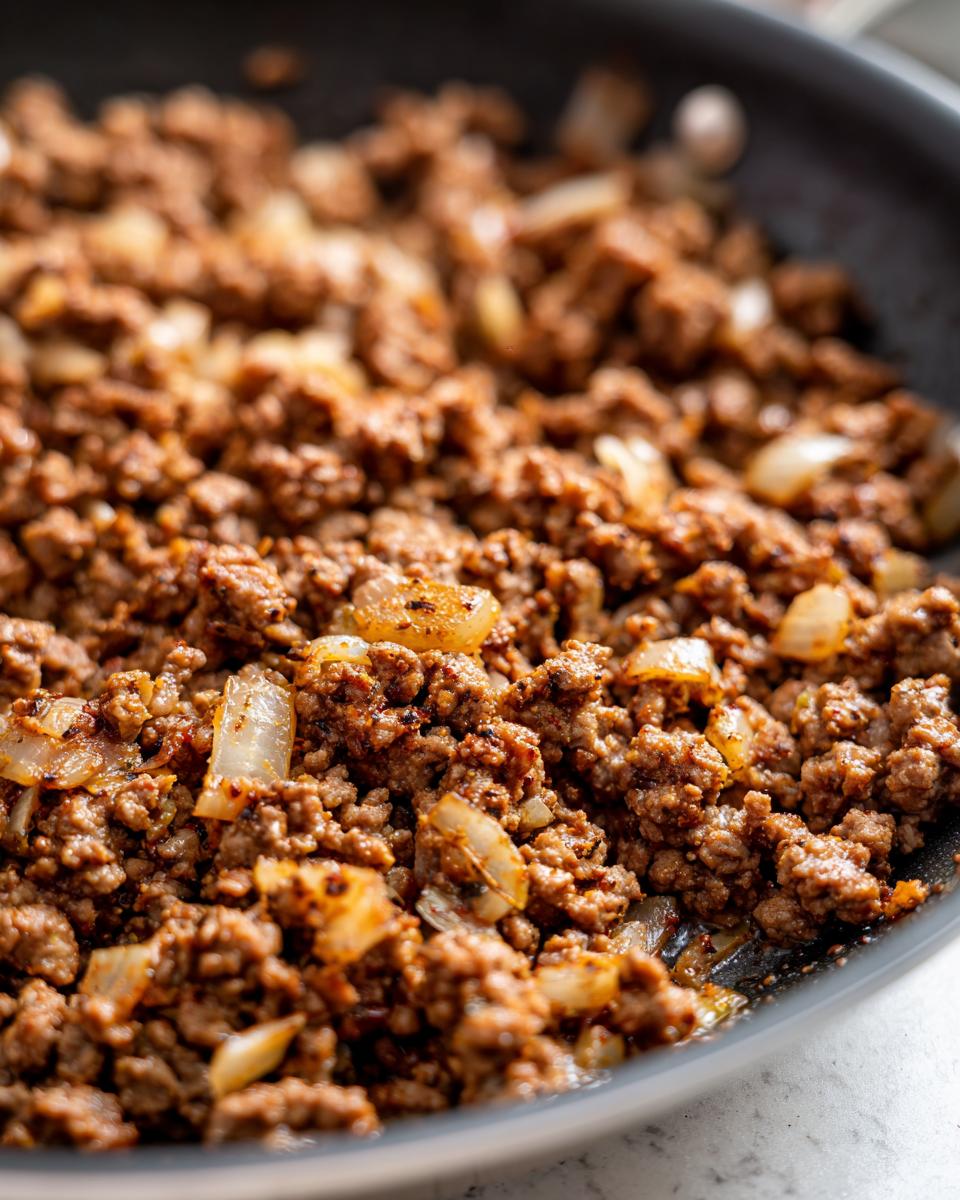 Close-up of cooked ground beef with sautéed onions in a black skillet for healthy ground beef recipes.