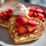 Heart-shaped valentines breakfast pancakes topped with sliced strawberries and whipped cream on a plate.