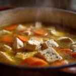 Close-up of steaming chicken soup with chunks of chicken and sliced carrots in broth.