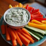 Plate of colorful carrot, cucumber, and bell pepper sticks with creamy herb dip for April spring snacks