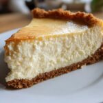 Close-up of a creamy cheesecake slice with a golden top and graham cracker crust on a white plate.