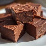 Close-up of creamy chocolate fudge squares stacked on a white plate, showcasing rich texture.