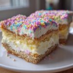Two creamy Easter dessert bars topped with white frosting and colorful round sprinkles on a white plate.