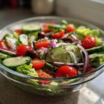 Glass bowl of fresh Greek salad with cucumbers, cherry tomatoes, red onions, olives, and lettuce.
