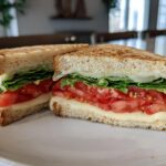 Grilled sandwich with melted cheese, fresh tomato slices, and leafy lettuce on white bread.