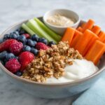 Bowl of healthy snack recipes featuring yogurt, granola, berries, celery, carrots, and dip.