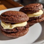 Two mini brownie cookie sandwiches with creamy filling on a white plate.