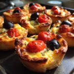 Close-up of mini savory tarts topped with cherry tomatoes and black olives on a baking tray, easy party snacks.