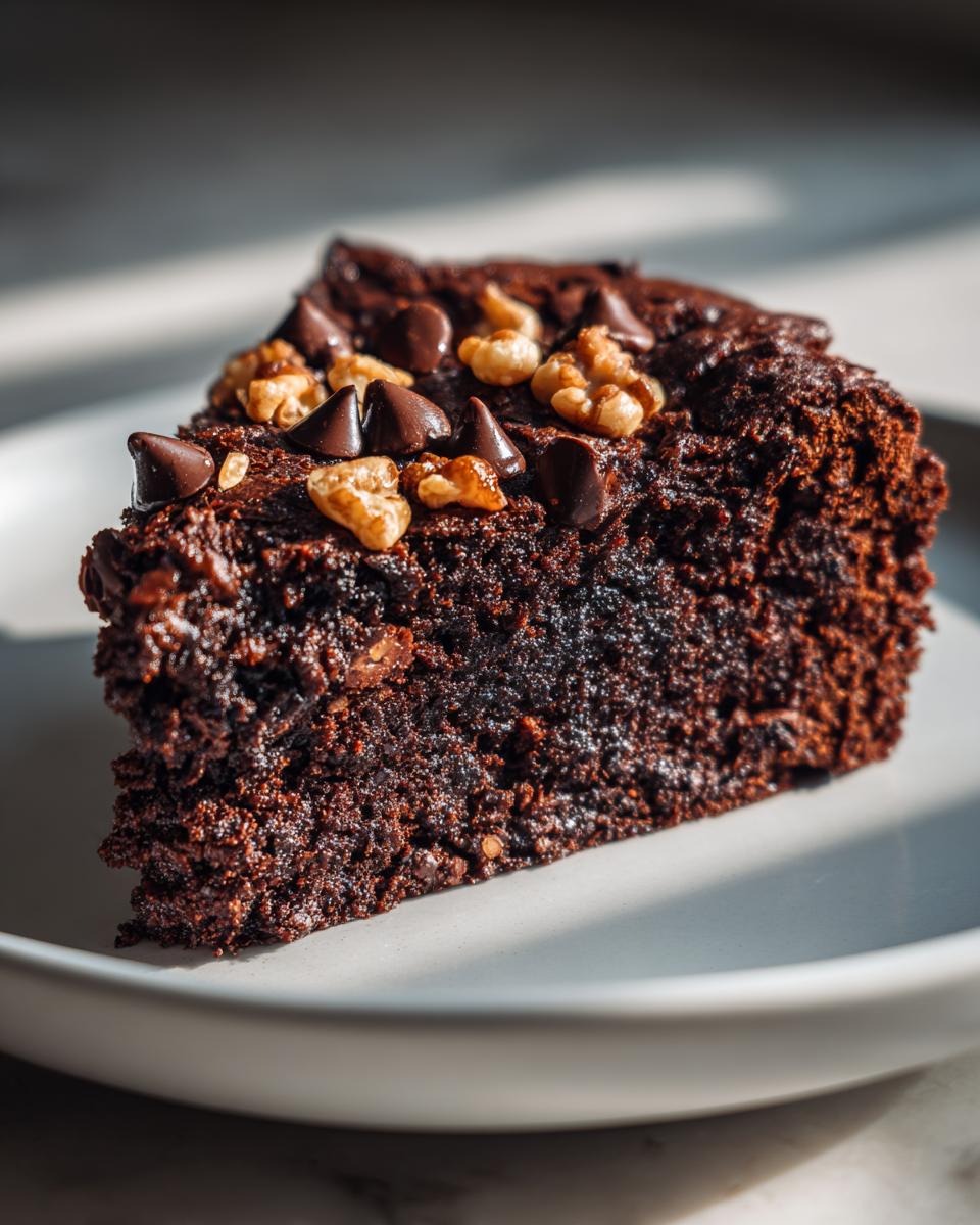 Close-up of a moist chocolate cake slice topped with walnuts and chocolate chips on a white plate.