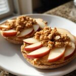 Bagel halves topped with peanut butter, apple slices, and granola on a white plate