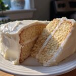 Slice of classic vanilla layer cake with white frosting on a white plate, perfect April holiday desserts